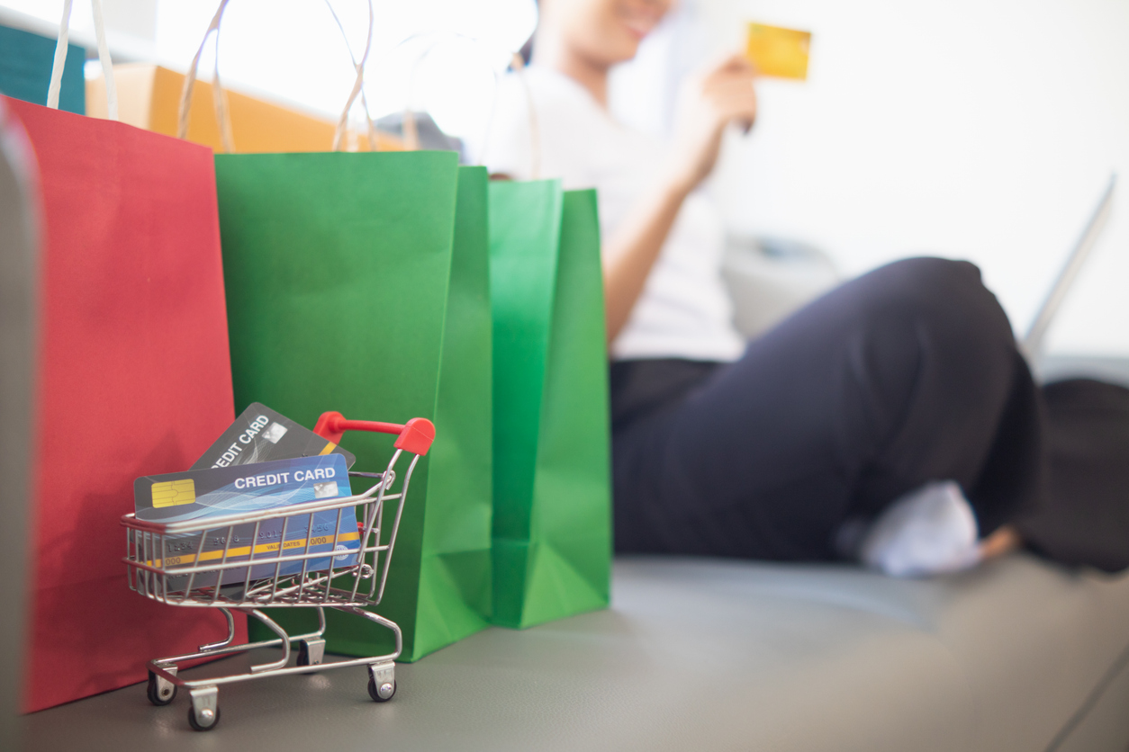 Credit cards in small shopping cart next to red and green shopping bags and a woman on a sofa holding a credit card